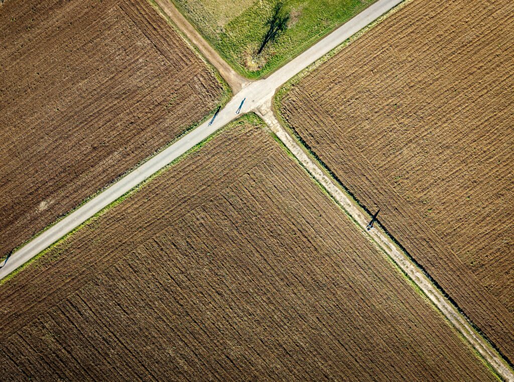 Overhead view of intersecting rural roads forming an X-shape between large brown plowed fields, with a small patch of green grass and a tree at the intersection.
